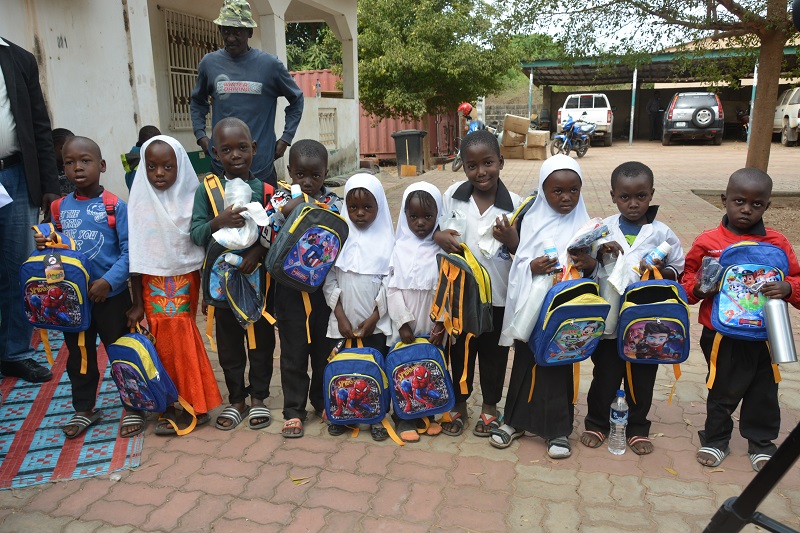Children smile at the camera in front of a school holding backpacks
