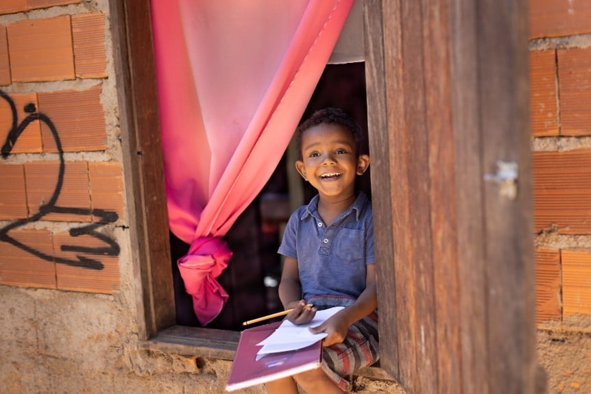A cheerful young boy sits in a wooden window frame, holding a notebook and pencil, with a pink curtain tied back beside him.