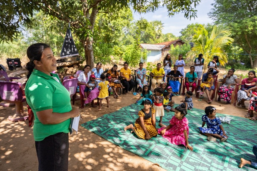 An aid worker talks to a group of parents surounding their children sitting on a blanket.