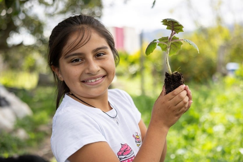 A smiling girl with dark hair holds a small seedling with soil, standing in a green outdoor environment.