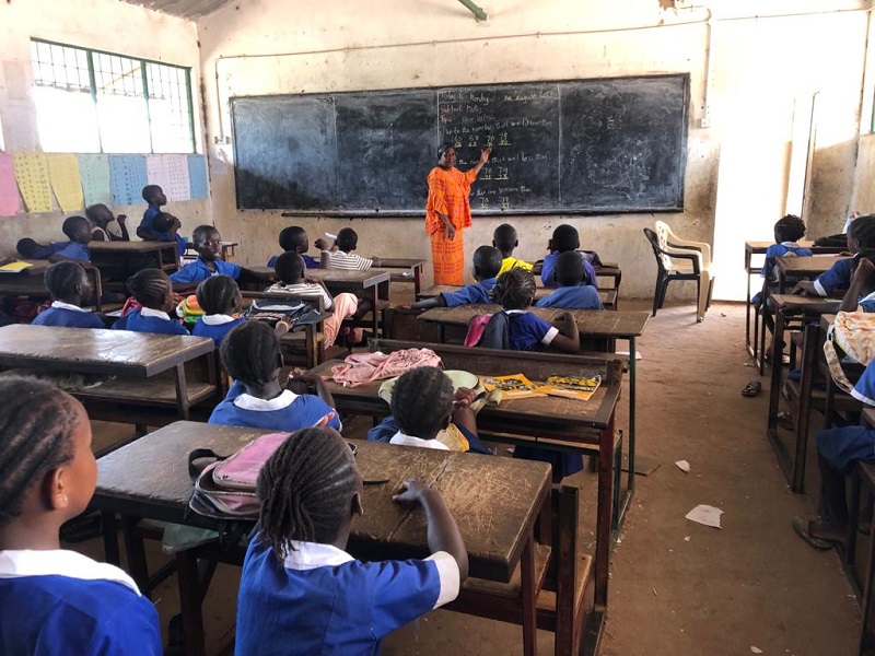 A group of children sit in a classroom in The Gambia as a teacher stands in front of a blackboard.