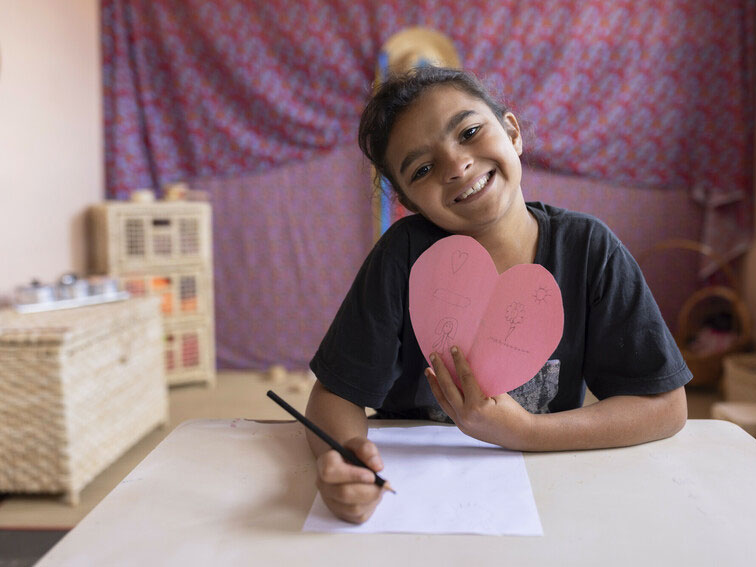 A smiling girl sits at a table, holding a pink heart-shaped drawing while holding a pencil, with a cozy, decorated indoor setting in the background.