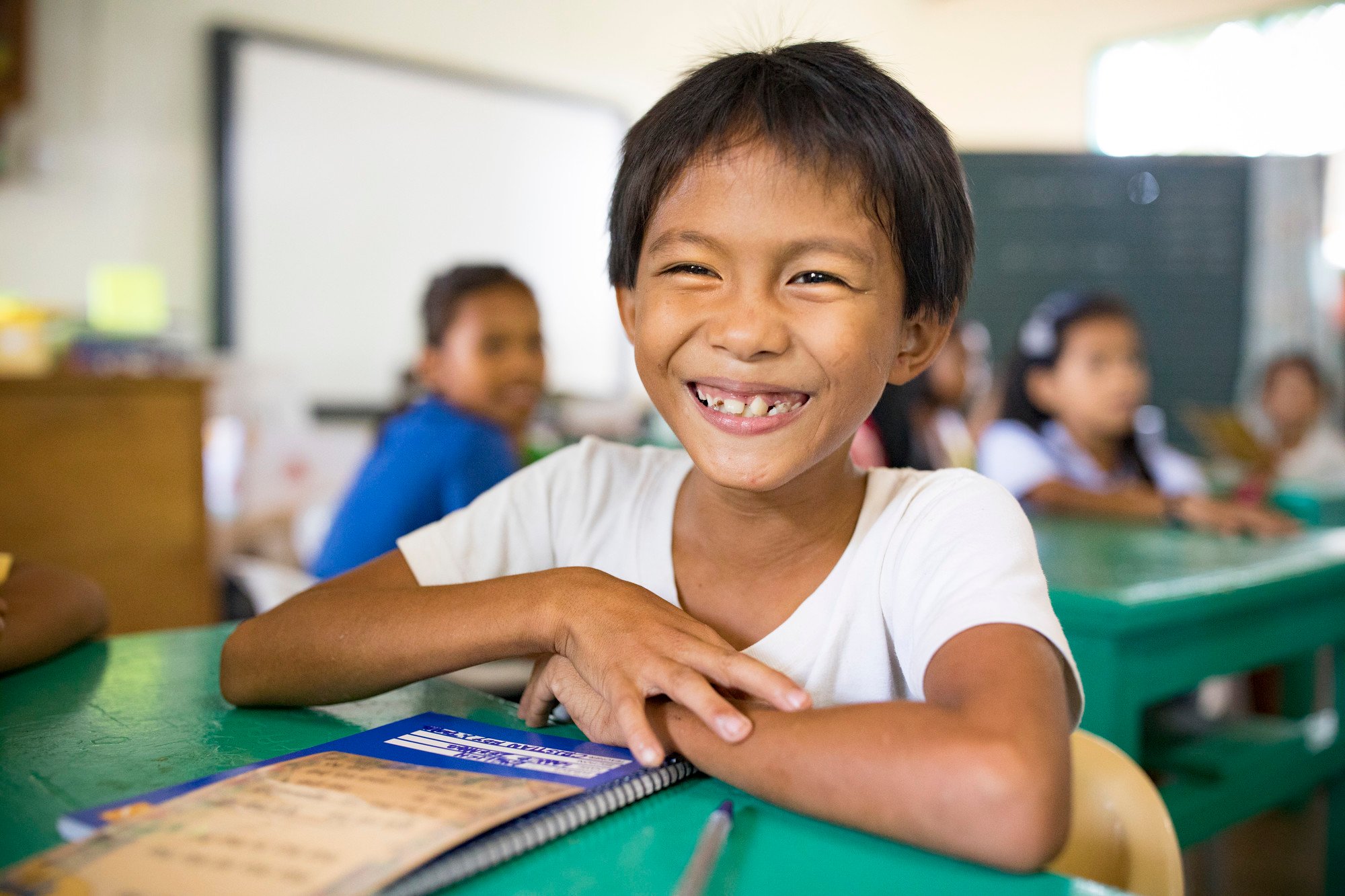 Smiling student sitting at a green desk in a classroom with other children studying in the background.