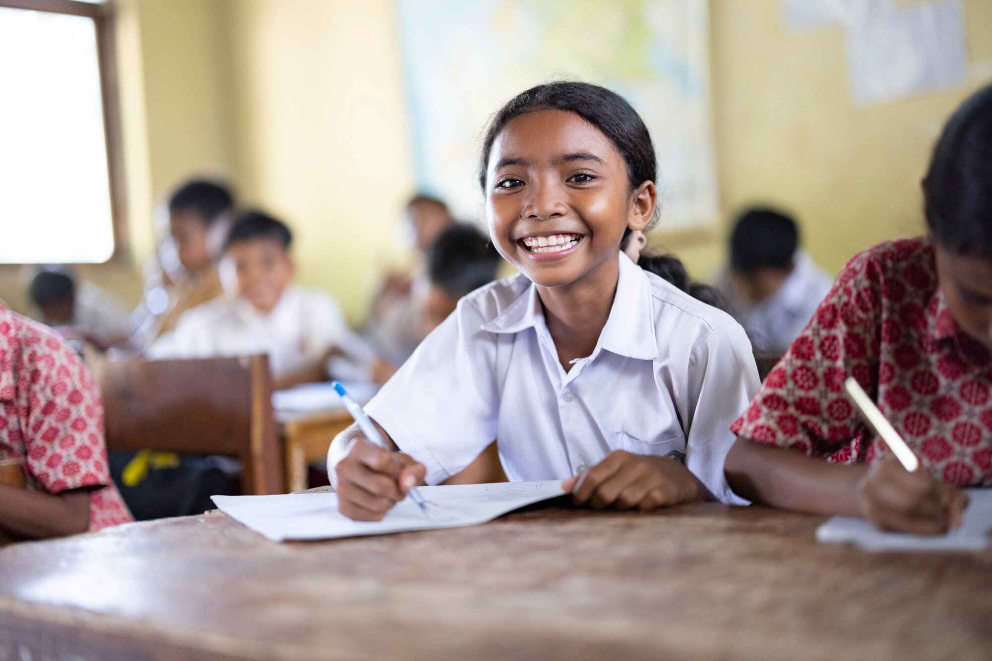 A girl in Indonesia sits at a desk in a classroom, smiling brightly.