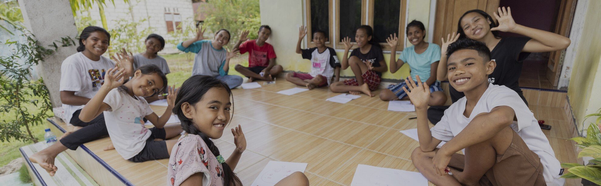 A group of children and young people sit together on a tiled outdoor area, smiling and waving at the camera while participating in a learning session.
