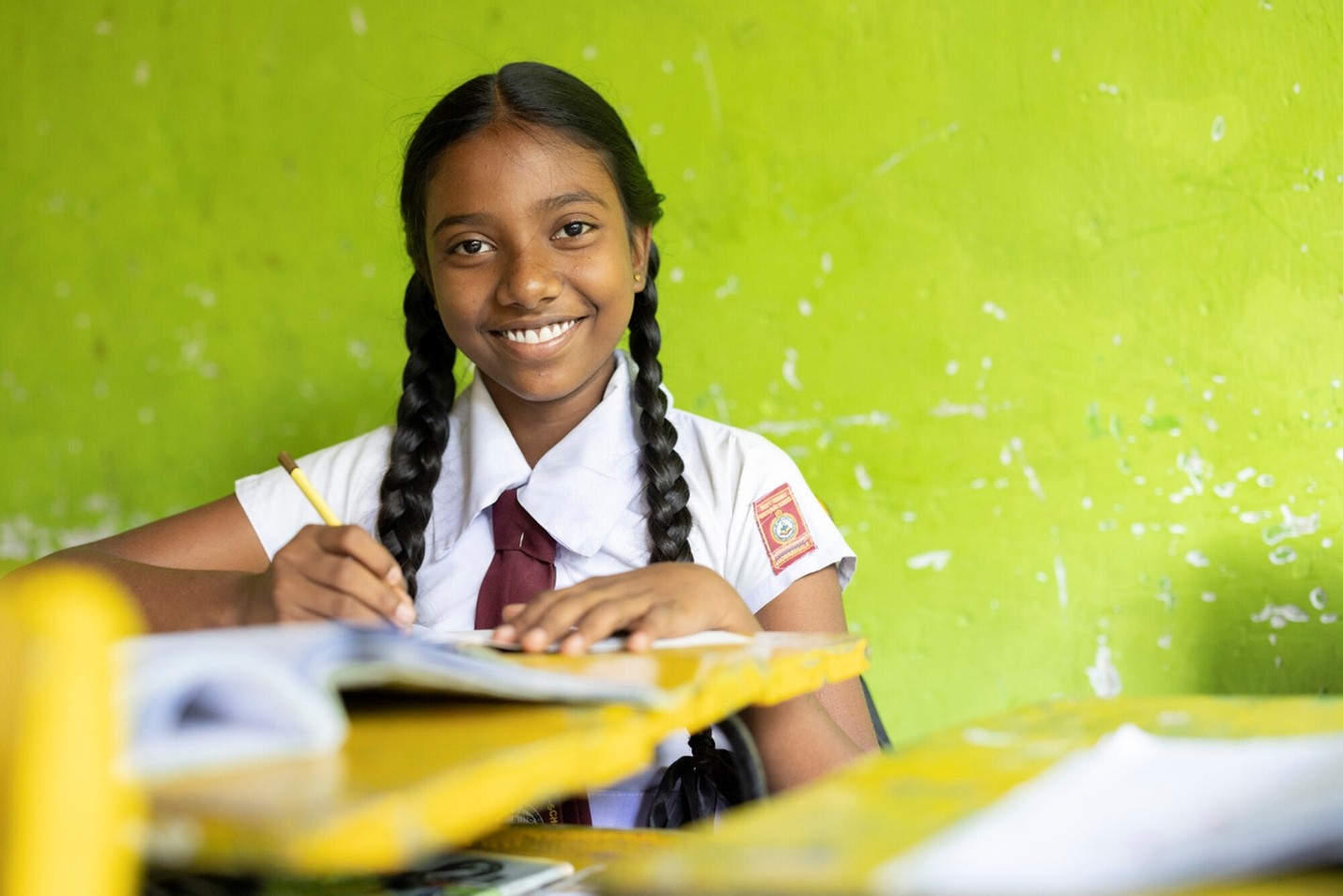 A smiling girl in a white school uniform with a red tie sits at a yellow desk, writing in her notebook, with a bright green wall in the background.