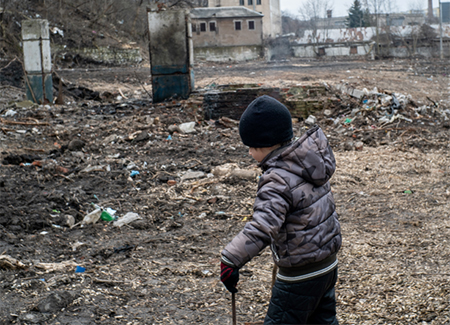 A child in a winter jacket and hat walks through a debris-strewn, war-torn area with damaged buildings in the background.