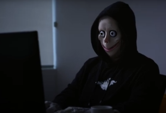 A person wearing a creepy mask and a hood sits at a desktop computer.